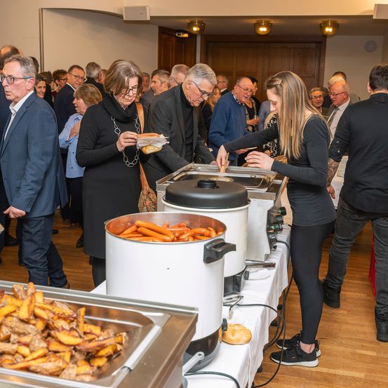 In einem Innenraum versammeln sich mehrere Personen um einen Tisch. Eine Frau serviert Essen, und andere holen sich ihre Teller. Es gibt Tabletts mit Hot Dogs, Brot und Pommes Frites. Der Raum ist gut beleuchtet, und es gibt Holzböden und eine Holztür.