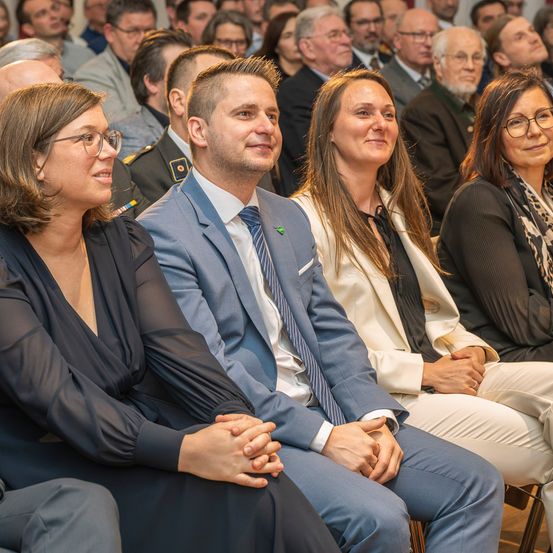 Eine Gruppe Erwachsener in formeller Kleidung sitzt in Reihen bei einer Veranstaltung, lächelt und blickt nach vorne, mit einem Mann in Uniform dahinter.