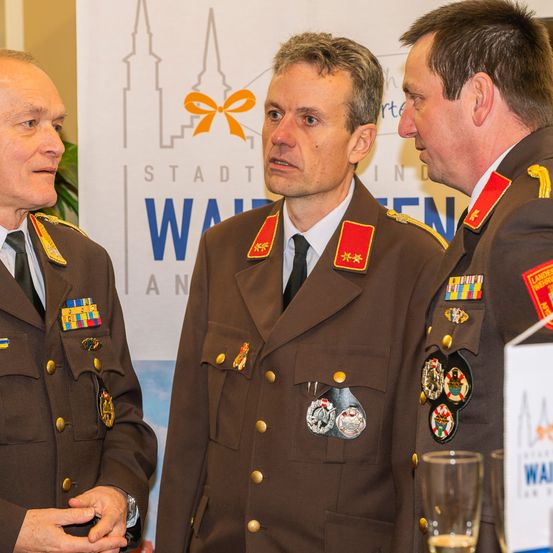 Drei Männer in Militäruniformen stehen vor einem Banner. Der Mann in der Mitte sieht ernst aus, während die anderen beiden ihn anschauen.