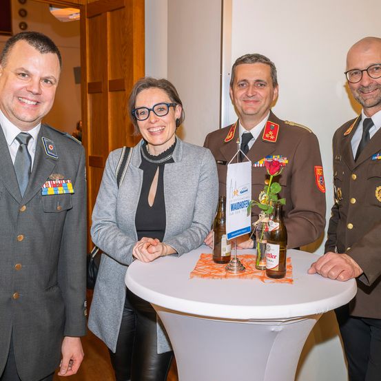 Vier Personen in Militäruniformen posieren für ein Foto; eine Frau lächelt, ein Mann hält eine Rose und zwei Männer stehen auf beiden Seiten mit einem Tisch in der Mitte.