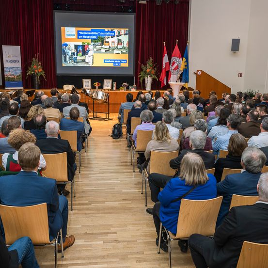 Ein Konferenzraum mit einer großen Leinwand, die eine Präsentation über die Stadt Waidhofen zeigt. Viele Menschen sitzen in Reihen und hören aufmerksam zu. Flaggen und Banner sind auf der Bühne zu sehen.