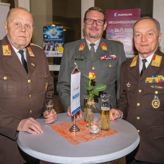 Drei Männer in Militäruniformen stehen um einen Tisch, lächeln. Zwei halten Gläser, einer von ihnen hat eine Rose und eine Flagge. Der Mann auf der rechten Seite trägt eine Medaille. Ein Plakat mit 'NEUJAHRSKONZERT' steht hinter ihnen.