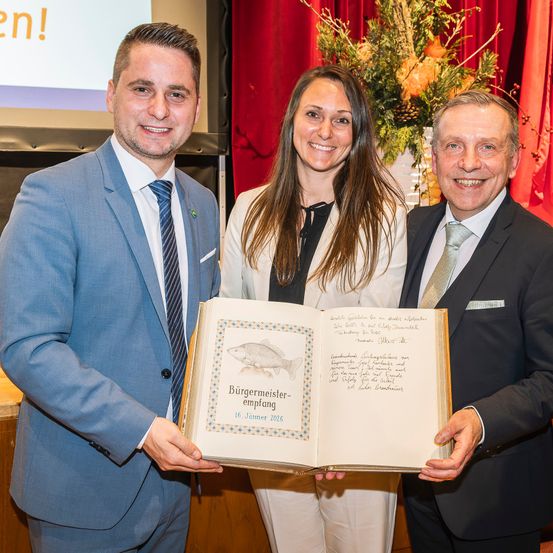 Drei Personen posieren für ein Foto, während sie ein offenes Buch mit dem Titel Burgermeister Empfang auf einem goldenen Hintergrund halten.