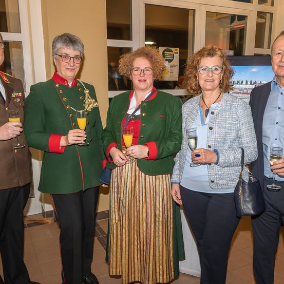 Fünf Personen stehen zusammen und halten Gläser mit Orangensaft und Wein. Sie sind in verschiedenen Kleidungsstücken gekleidet, einige in Uniformen. Hinter ihnen zeigt ein großes Fenster eine Ansicht von Gebäuden.
