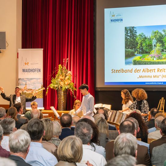 Ein großes Publikum beobachtet, wie ein Mann vor einer Projektionsleinwand, die ein Parkbild zeigt, auf der Bühne Trommeln spielt. Im Vordergrund interagieren zwei Personen mit Metallinstrumenten.