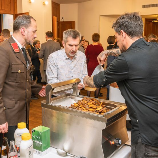 Ein Mann in Uniform spricht mit einem Mann in einem weißen Hemd. Ein Koch serviert Essen von einem Buffet an einen Mann in einem schwarzen T-Shirt.