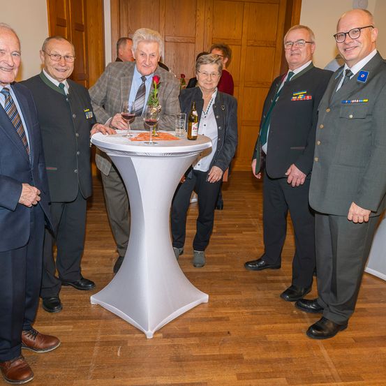 Eine Gruppe älterer Männer und Frauen in formeller Kleidung steht in der Nähe eines Tisches mit Weingläsern und einer Flasche, möglicherweise bei einer formellen Veranstaltung.