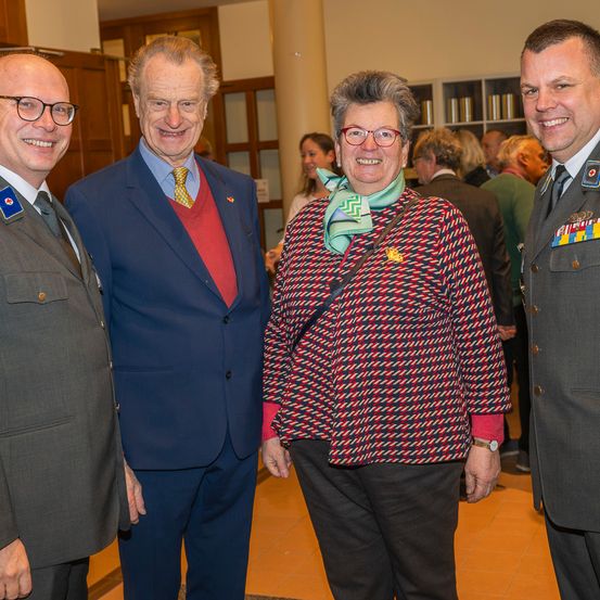 Vier Menschen in formeller Kleidung stehen zusammen und lächeln für ein Foto. Eine Frau in der Mitte trägt eine Krawatte und eine Brille. Hinter ihnen stehen und sprechen Menschen.