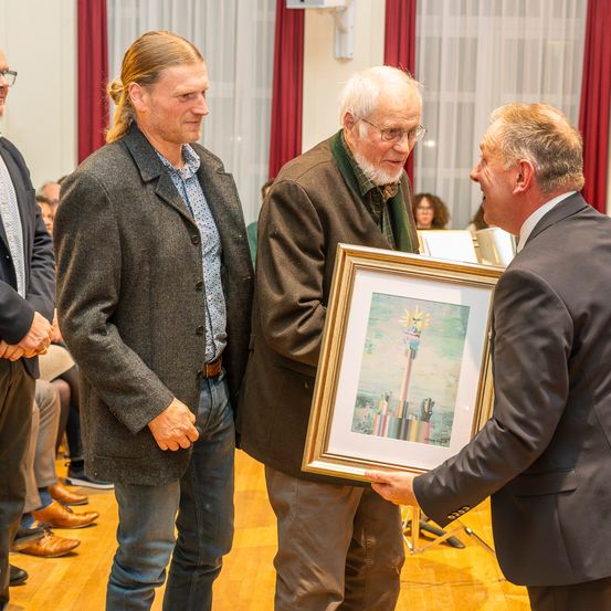 Drei Männer stehen drinnen, einer hält ein gerahmtes Bild. Der Mann auf der linken Seite trägt einen Mantel und Jeans, während der Mann in der Mitte eine Brille und einen Pullover trägt. Der Mann auf der rechten Seite trägt einen Anzug und hält den Rahmen.