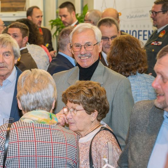 Eine Versammlung von Menschen in einem Raum, wahrscheinlich bei einer Veranstaltung. Ein Mann in einem Anzug und mit Brille steht in der Mitte und lächelt. Einige Leute schauen ihn an. Ein Schild mit Text ist im Hintergrund sichtbar.