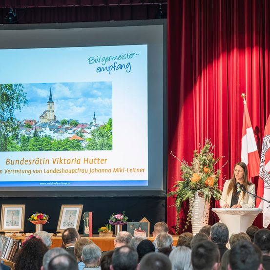 Eine Frau spricht an einem Podium mit einem Mikrofon vor einem Publikum. Ein großer Bildschirm hinter ihr zeigt eine Stadt und den Text 'Bürgermeister Empfang'. Ein großer roter Vorhang und Flaggen sind rechts.