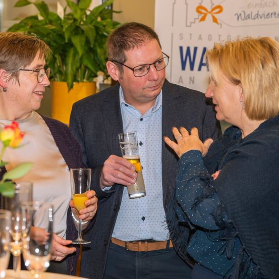 Drei Erwachsene, zwei Frauen und ein Mann, stehen in einer sozialen Umgebung. Der Mann hält ein Glas Bier und die Frauen halten Weingläser. Sie scheinen in ein Gespräch vertieft zu sein.