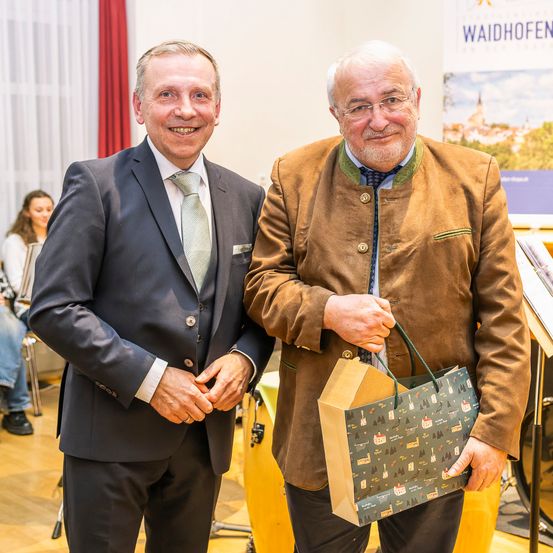 Zwei Männer stehen lächelnd nebeneinander, einer in Anzug und der andere in einer braunen Jacke, und halten eine Geschenktüte. Sie sind in einem Raum mit Holzböden und einem Banner im Hintergrund.
