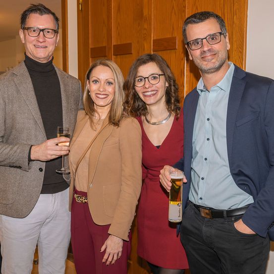 Vier Menschen stehen zusammen, zwei Frauen und zwei Männer, lächeln und posieren für ein Foto. Der Mann auf der linken Seite hält ein Glas Champagner. Der Mann auf der rechten Seite hält ein Glas Bier. Die Frauen tragen eine Brille und sind in elegante Kleidung gekleidet. Hinter ihnen ist eine Holzwand.
