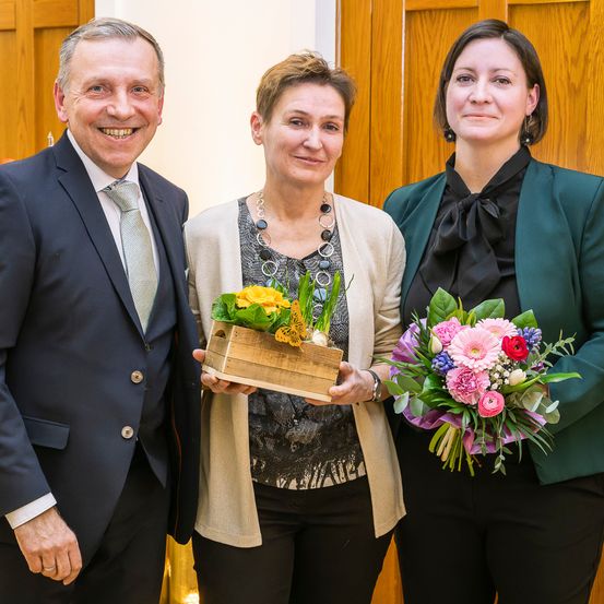 Drei Personen stehen zusammen, lächeln und posieren für ein Foto. Die Frau in der Mitte hält eine Holzkiste mit Blumen und die Frau auf der rechten Seite hält einen Blumenstrauß. Sie sind alle formell gekleidet.