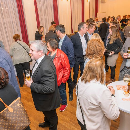 Eine Gruppe von Menschen versammelt sich in einem Raum, vielleicht für eine soziale Veranstaltung. Einige stehen und andere sitzen. Einige halten Getränke und andere führen Gespräche. Es gibt einen Tisch mit Flaschen und Gläsern darauf.