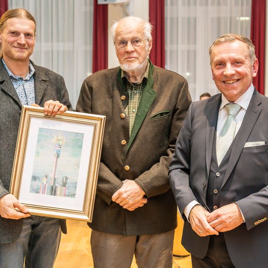 Drei Männer stehen zusammen in einem Raum. Der Mann auf der linken Seite hält ein gerahmtes Bild. Der Mann in der Mitte trägt eine Brille und eine braune Jacke. Der Mann auf der rechten Seite trägt einen Anzug und eine Krawatte.