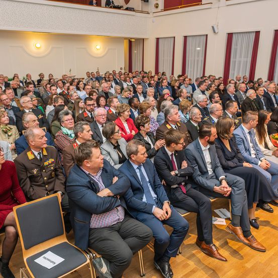 Ein großer Saal mit Menschen in Reihen, möglicherweise bei einer formellen Veranstaltung. Einige sind in Militäruniformen, andere in Anzügen. Der Raum ist von warmen Lichtern beleuchtet, und eine Frau in einem roten Kleid sitzt vorne.