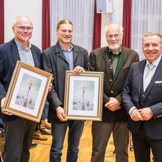 Vier Männer posieren für ein Foto und halten gerahmte Kunstwerke in den Händen. Sie stehen in einem Raum mit Holzböden und roten Vorhängen. Die Männer tragen formelle Kleidung.