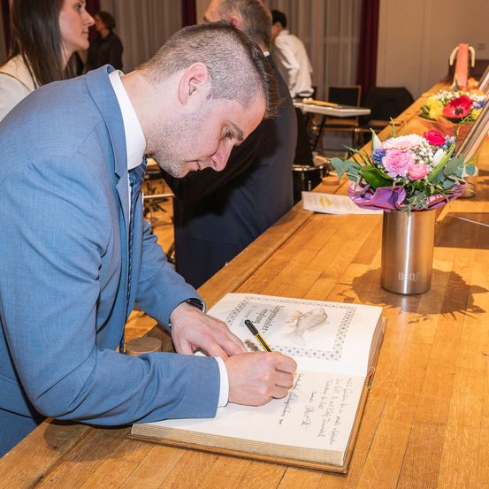 Ein Mann in einem blauen Anzug unterschreibt ein Buch auf einem Holztisch mit Blumen in einer Vase in der Nähe. Hinter ihm stehen Menschen und schauen auf das Buch.
