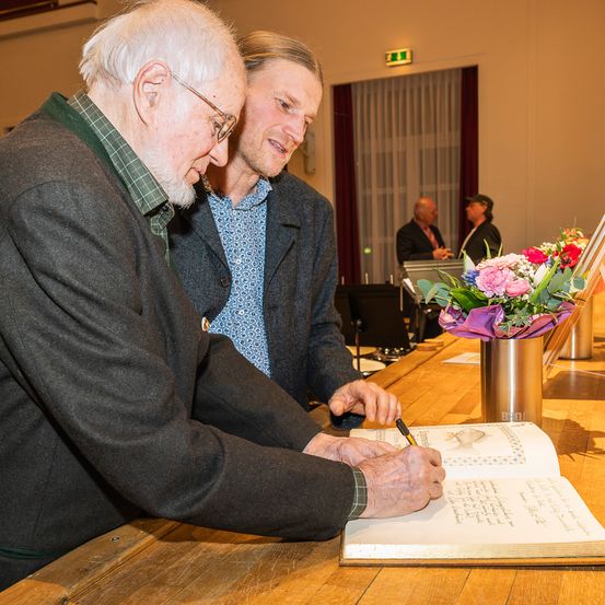 Ein älterer Mann unterzeichnet ein Buch mit einem Stift an einem Tisch. Neben ihm blickt ein jüngerer Mann zu. Ein Blumenarrangement und ein Mikrofon sind auf dem Tisch. Im Hintergrund unterhalten sich zwei Personen in der Nähe einer weißen Gardine.