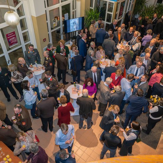 Eine Indoor-Versammlung verschiedener Personen, möglicherweise bei einer formellen Veranstaltung, wobei einige in Militäruniformen sind. Sie stehen um Tische herum, trinken und unterhalten sich. Im Hintergrund befindet sich ein Fernsehbildschirm.