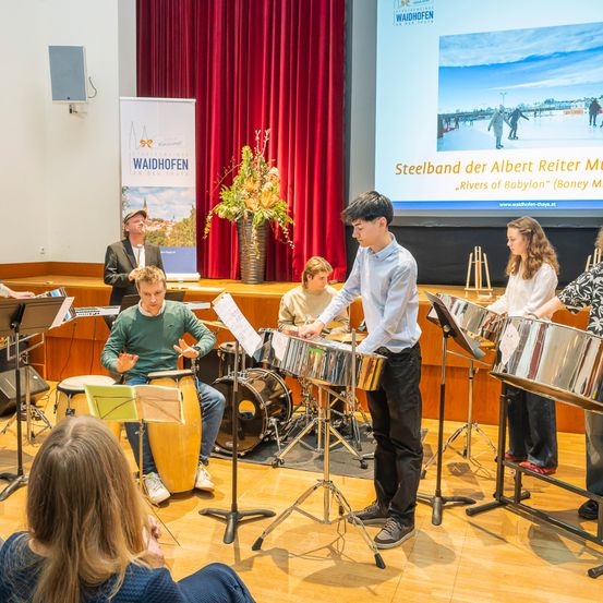 Eine Gruppe von Musikern spielt Steel Drums und andere Percussionsinstrumente auf einer Bühne in einem Klassenzimmer. Eine Beamerleinwand zeigt den Titel Steelband der Albert Reiter Music und die Komposition Rivers of Babylon.