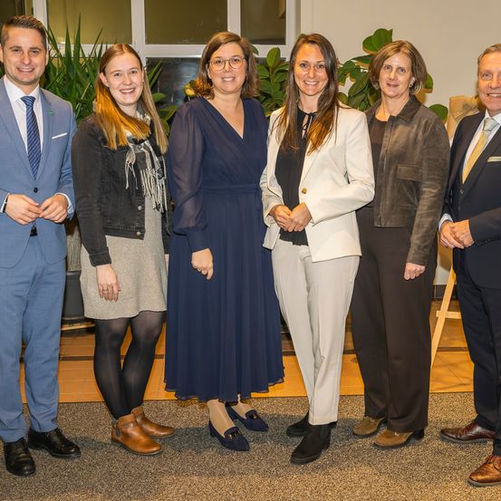 Eine Gruppe von Menschen, die feierlich gekleidet sind, stehen zusammen in einem Raum, lächeln und posieren für ein Foto. Es gibt drei Frauen und drei Männer, mit Pflanzen im Hintergrund.