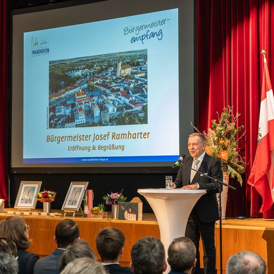 Ein Mann steht an einem Rednerpult mit Mikrofon und präsentiert vor einem Publikum. Eine große Leinwand zeigt eine Stadtlandschaft und den Text 'Bürgermeister Josef Ramharter Eröffnung & Begrüßung'. Eine Flagge steht hinter dem Redner.