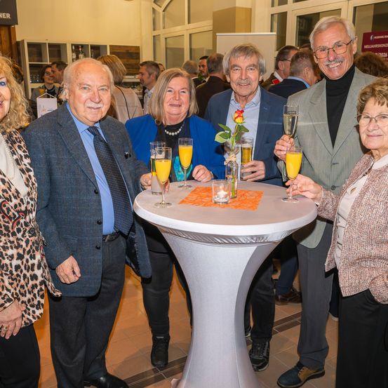 Eine Gruppe von Menschen steht um einen Tisch mit Getränken und Blumen. Sie lächeln und scheinen eine Feier zu veranstalten. Der Tisch hat Gläser mit Orangensaft und einen Blumenstrauß. Im Hintergrund sind andere Menschen zu sehen, die möglicherweise an derselben Veranstaltung teilnehmen.