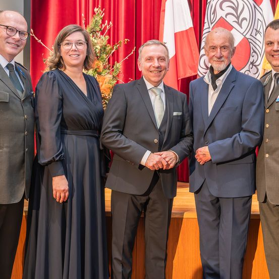 Fünf Personen stehen vor einer Bühne mit Fahnen im Hintergrund. Drei Männer und zwei Frauen, alle in Anzügen, lächeln für die Kamera.