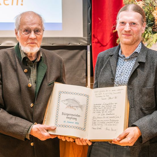 Zwei Männer stehen und halten ein Buch mit dem Titel 'Bürgermeister-Empfang' und einem Datum vom 16. Januar 2016. Sie lächeln beide und schauen nach vorne. Hinter ihnen ist ein roter Vorhang. Der Mann auf der linken Seite trägt eine Brille und eine geknöpfte Jacke. Der Mann auf der rechten Seite trägt ein gemustertes Hemd und eine Jacke.