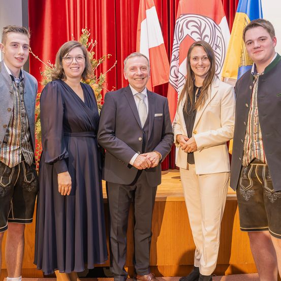Fünf Personen stehen vor einer Bühne, gekleidet in formelle Kleidung. Drei Männer und zwei Frauen tragen traditionelle deutsche Kleidung. Hinter ihnen befinden sich drei Flaggen auf der Bühne und ein Weihnachtsbaum auf der linken Seite.