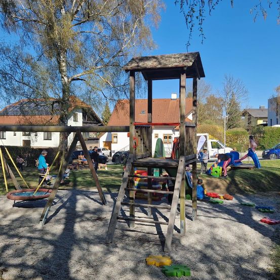 Ein Spielplatz im Freien mit Schaukeln, Rutschen und Klettergerüsten. Einige Kinder spielen und Erwachsene sind anwesend. Bäume und Häuser sind im Hintergrund.