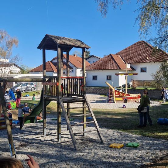 Ein Spielplatz mit Rutsche und Leiter, umgeben von einem roten Boot und mehreren Häusern. Leute spielen und einige gehen.