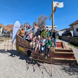 Eine Gruppe von Menschen, Erwachsene und Kinder, steht auf und um ein großes Piratenschiff-Spielplatz herum. Das Schiff hat eine Piratenflagge oben und steht in einem Park.