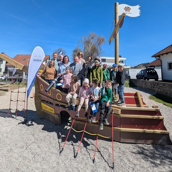 Eine Gruppe von Menschen, Erwachsene und Kinder, steht auf und um ein großes Piratenschiff-Spielplatz herum. Das Schiff hat eine Piratenflagge oben und steht in einem Park.