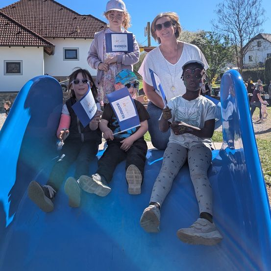 Fünf Kinder sitzen lächelnd auf einem blauen Rutschvergnügen. Sie halten blaue Fahnen und ein Kind trägt einen Hut. Hinter ihnen steht eine Frau und hält eine blaue Fahne. Im Hintergrund befindet sich ein Haus und einige Menschen.