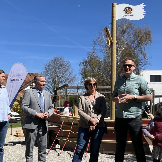 Fünf Personen stehen im Freien. Ein Mann in Anzug und Krawatte steht neben einer Frau im Blazer. Dahinter ist ein Fahnenmast mit einer Piratenflagge und ein Schild zu sehen. Ein Kind mit rotem Hut steht in der Nähe eines Spielplatzes.