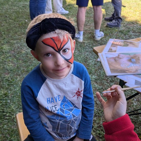 Ein Junge mit einem Superhelden-Gesichtsgemälde sitzt auf einer Bank, während andere im Hintergrund stehen.