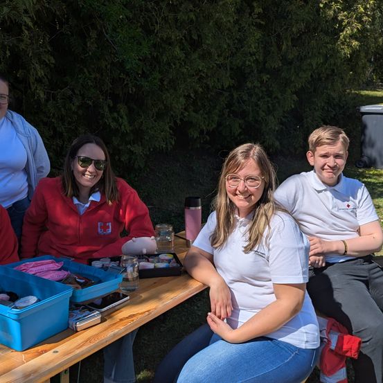 Vier Personen sitzen um einen Picknicktisch mit Snacks. Eine Frau trägt eine rote Jacke. Eine Frau in einem weißen T-Shirt lächelt. Ein Junge in einem weißen Polohemd sitzt neben ihr.