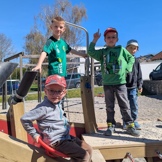 Vier Jungen sind auf einem Spielplatz. Ein Junge sitzt auf einem roten Schaukelpferd. Die anderen stehen um ihn herum. Dahinter steht ein Gebäude und geparkte Autos.