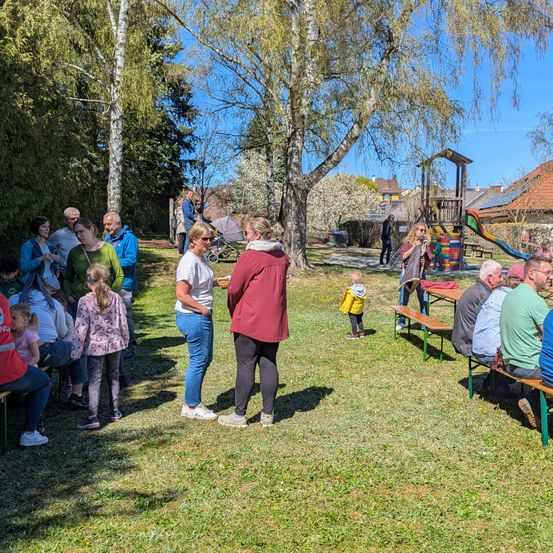 Eine Gruppe von Menschen steht und sitzt in einem Grasbereich mit Bäumen und einem Spielplatz. Eine Frau spricht zu einer Gruppe von Menschen.