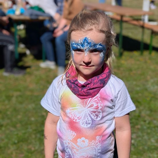 Ein kleines Mädchen steht auf einem Rasenfeld und trägt ein weißes T-Shirt mit bunten Schmetterlingsmustern und einem roten Schal. Sie hat blaue Gesichtsbemalung mit Schneeflocken auf dem Gesicht.
