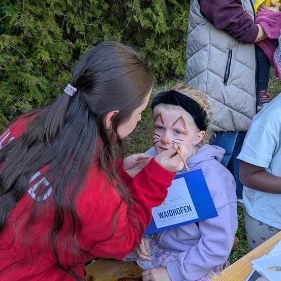 Eine Frau malt Katzenohren auf das Gesicht eines jungen Mädchens. Eine andere Frau hält ein Schild mit 'Waidhofen'. Sie sitzen auf einer Bank. Dahinter hält ein Mann ein Kind.
