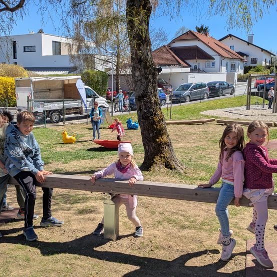 Mehrere Kinder spielen in einem Park. Ein Mädchen lehnt sich an eine Wippe mit einem anderen Mädchen, während zwei weitere Kinder auf der Wippe sitzen. Eine Frau steht in der Nähe, und verschiedene Spielzeuge und Häuser sind im Hintergrund.