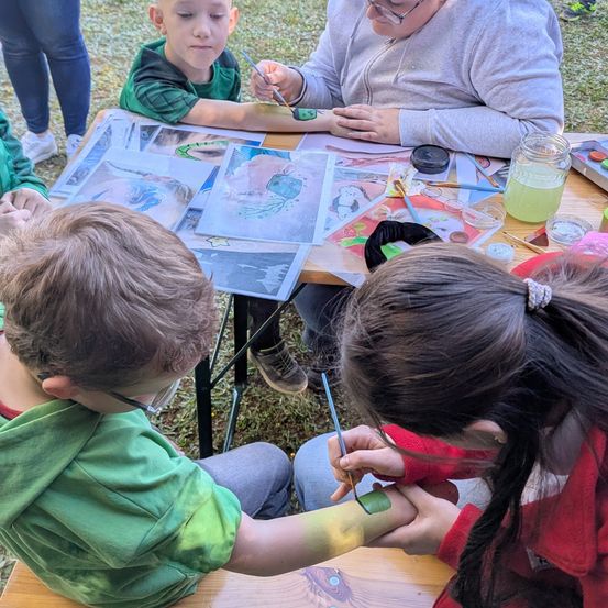 Kinder und Erwachsene malen auf einem Tisch im Freien. Ein Kind trägt eine Brille und ein grünes Shirt. Eine erwachsene Frau hilft einem anderen Kind, etwas auf seinen Arm zu malen.