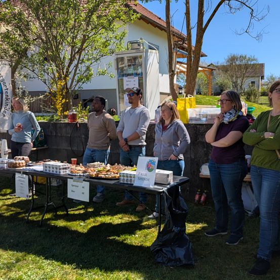 Eine Gruppe von Menschen steht an einem mit Essen gefüllten Tisch an einem sonnigen Tag. Der Tisch hat Schilder für Getränke und Essen. Die Leute schauen sich das Essen an.