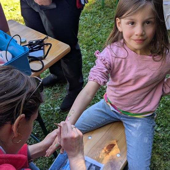 Ein Mädchen lässt sich von einer Frau in einer roten Jacke eine Blumenzeichnung auf den Arm malen. Im Hintergrund steht ein Mann in der Nähe eines Holztisches mit verschiedenen Gegenständen darauf.