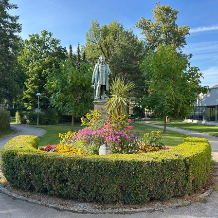 Eine Statue eines Mannes steht in der Mitte eines kreisförmigen Gartens mit bunten Blumen. Der Garten ist von einer Hecke umgeben und befindet sich in einem Park mit grünen Bäumen und einem klaren blauen Himmel.